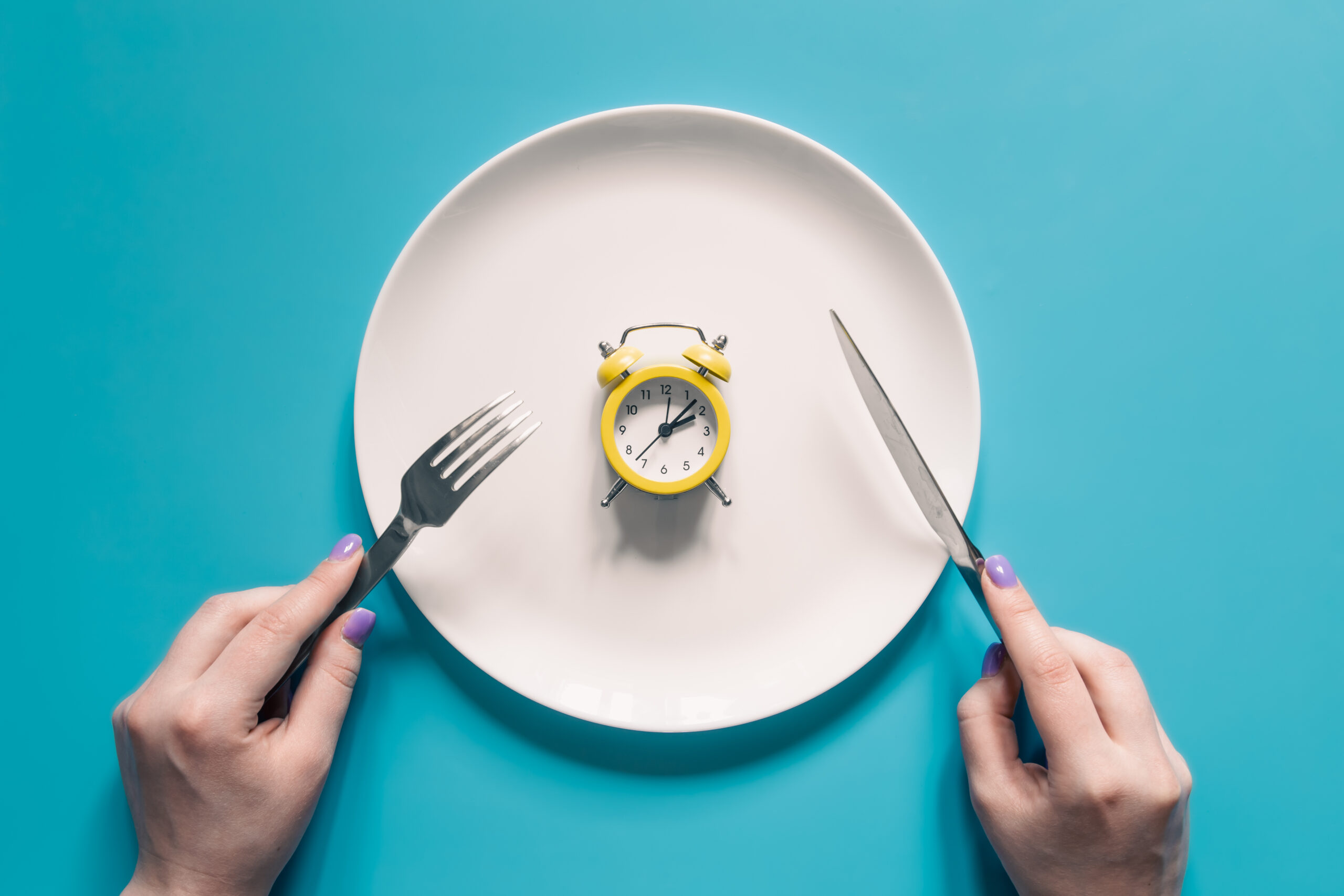 Clock on a dining table with an empty plate, fork, and knife, symbolizing time-restricted eating and intermittent fasting.