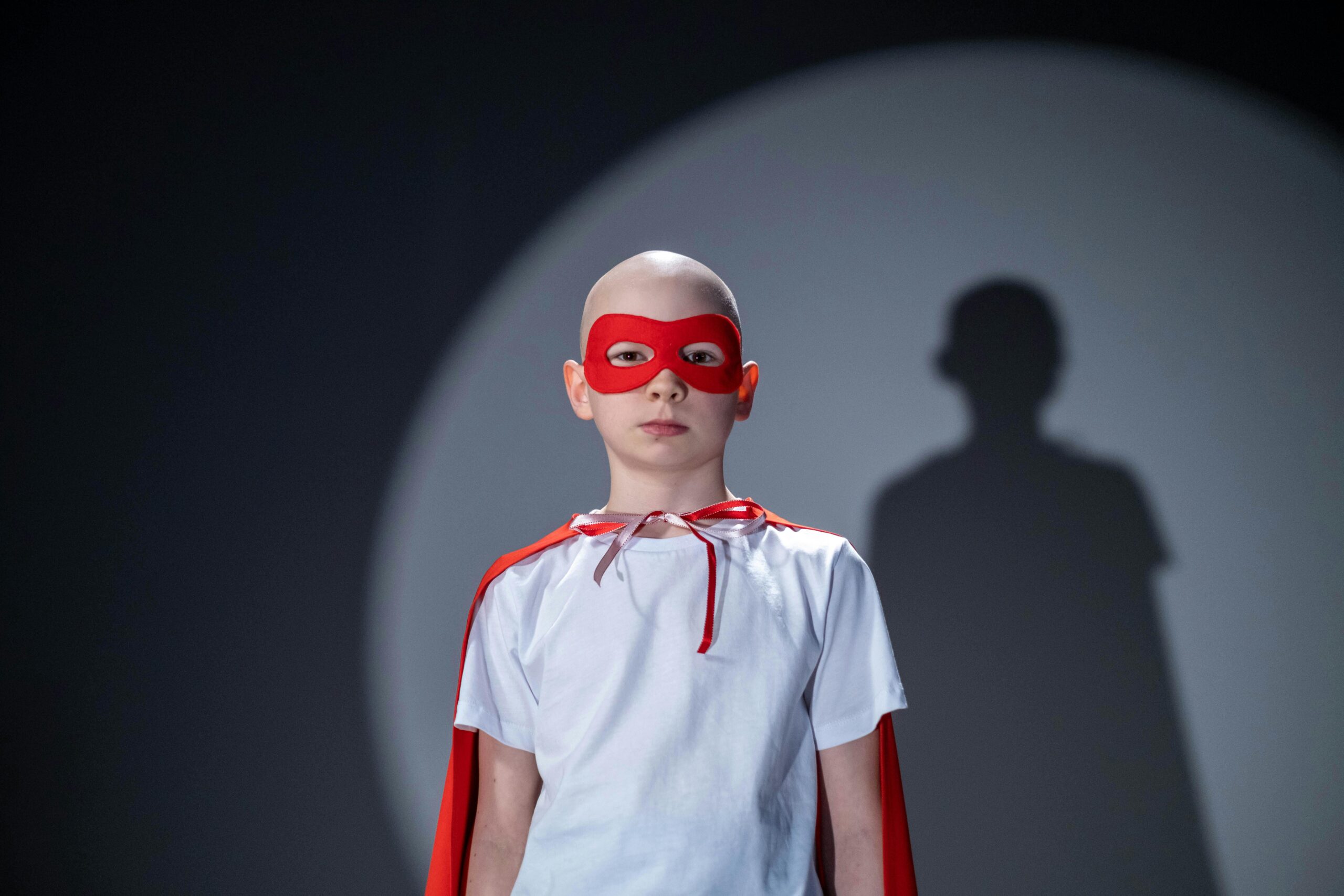Portrait of young superhero in a white shirt, red cape, and mask with a bold shadow behind.